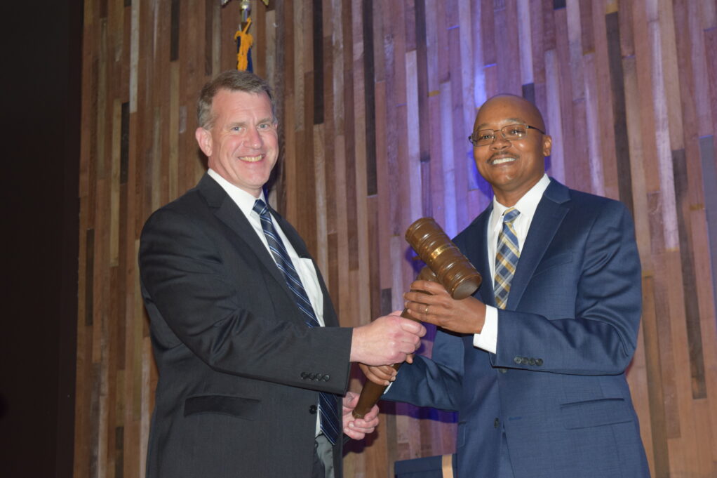 Jon Heyl and Clayton Morgan stand holding the gavel between them. Jon Heyl, a white man with brown hair who is wearing a navy blue and grey striped tie and black suit. Clayton Morgan is a Black man with brown hair, and he is wearing glasses, a dark blue suit, a white shirt, and a grey and white plaid tie. Jon and Clayton stand in front of a background with gold and silver streamers.