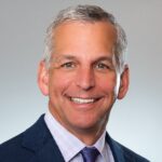 Alex Hagan is a white man with grey hair. he is pictured wearing a white shirt with a dark blue tie and a black jacket. He stands in front of a grey background and is pictured smiling.