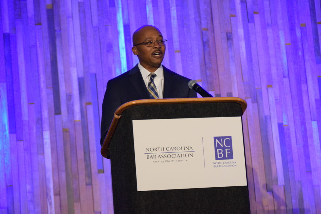 Clayton Morgan stands at a brown podium, which has a white banner with the NCBA logo on it. Clayton Morgan is a Black man with brown hair, and he is wearing glasses, a dark blue suit, a white shirt, and a grey and white plaid tie. He stands in front of a background with gold and silver streamers.