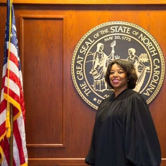 Judge Ola M. Lewis is a Black woman with dark brown hair. She is pictured wearing a black robe, and she is standing in front of a wood wall with the Great Seal of the State of North Carolina behind her, which is a round circle on the wall with gold letters and an image in gold, and the date May 20, 1775, on the seal, along with a silhouette of two people. The American flag hangs on a stand to the left of the photo. 