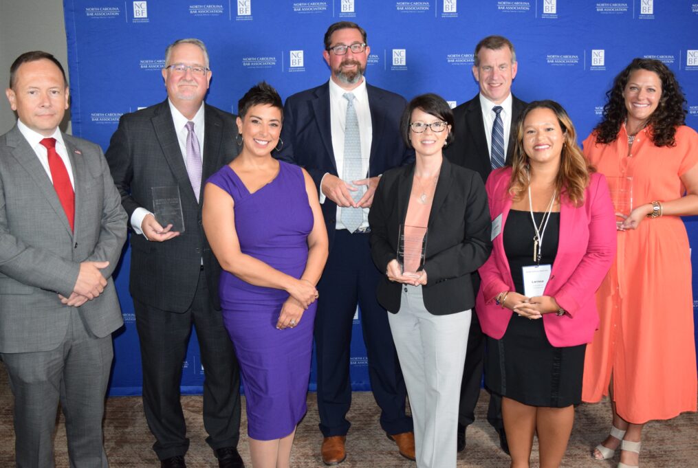 Eight individuals stand against a bright blue NCBA background. Front from left, are Michelle Loveys Dozier (Inner Banks Legal Services), Jackie Kiger (Pisgah Legal Services), and Larissa Mervin, Pro Bono Committee co-chair. Back: NCBA Executive Director Jason Hensley, Matt McGuire (Alston & Bird), Ben Finholt (Duke Law School), NCBA President Jon Heyl, and Sarah Beth Winters (Inner Banks Legal Services). Michelle Loveys Dozier is a white woman with short dark hair, and she is smiling and wearing a purple dress with gold hoop earrings. Jackie Kiger is a white woman with chin-length dark-brown hair and dark brown glasses, and she is smiling and wearing a pale pink blouse, black blazer, and pale grey pants. Larissa Mervin is a Hispanic woman with golden brown shoulder-length hair, and she is smiling and wearing a black dress and pink blazer. Jason Hensley is a white man with brown hair who is wearing a red tie and a grey suit with an American flag pin on his lapel. Matt McGuire is a white man with grey hair and a short grey beard, and he is smiling with his mouth closed and wearing thin wire-rimmed glasses, a white shirt, black suit, and lavender tie. Ben Finholt is a white man with dark hair and a brown and grey beard and mustache, and he is smiling and wearing black glasses, a white shirt, and a black suit. Jon Heyl is a white man with brown hair who is wearing a black suit and a navy-blue and grey striped tie. Sarah Beth Winters is a white woman with curly brown shoulder-length hair, and she is smiling and wearing a long button-down peach dress. 