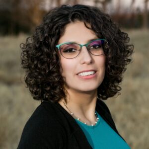 Yazmeen Gadalla is a white woman with curly dark brown hair. She is wearing teal glasses, a teal blouse, a black sweater, and she is pictured smiling.