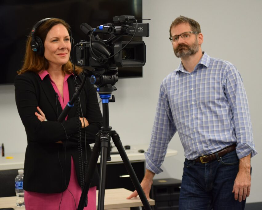 Ashley stands in the studio, in front of a camera with earphones on. She wears a black jacket and pink shirt and stands next to a white man with a beard in a plaid shirt.