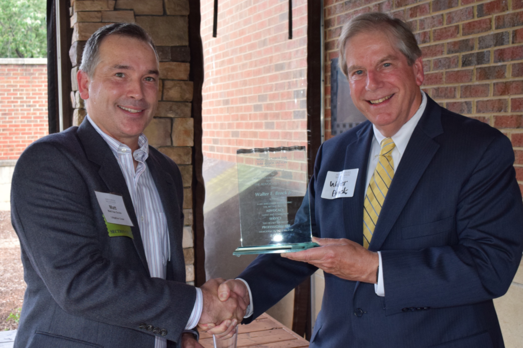 Matt, a white man with brown hair, wears a white and blue striped shirt and grey suit, and Walter, a white man with light brown hair, wears white shirt, yellow tie, and grey suit. Matt is handing the glass award to Walter.