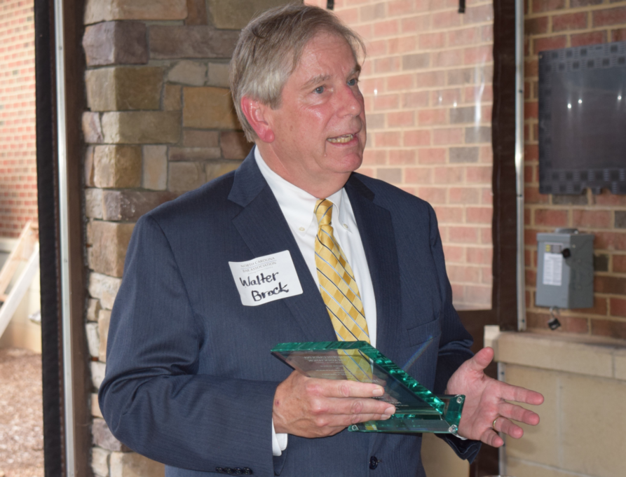 Walter, a white man with light brown hair, wears a white shirt, grey suit, and yellow tie and holds the advocate's award.