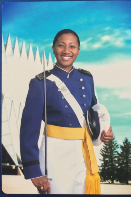 Jocelyn, a Black woman, is pictured as a cadet and wears a blue, white and gold uniform and white hat. Trees and sky are visible behind her.