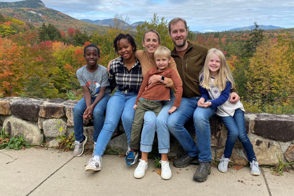 Dean is pictured sitting on a rock wall with his wife, his two daughters, and his two sons. Orange and green fall foliage is behind them.