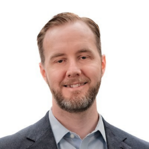 Jamie, a white man with brown hair and a beard, wears a pale grey shirt and dark grey suit and is smiling.