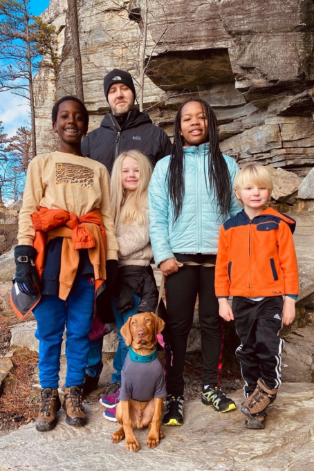 Dean, a white man with brown hair, wears a black jacket and hat. He, his four children, and their dog stand in front of a rock formation. 