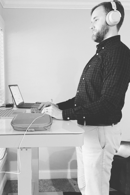 Dean is pictured in a black and white photo. He wears a dark shirt and khakis and headphones. He stands at a desk typing on a computer.