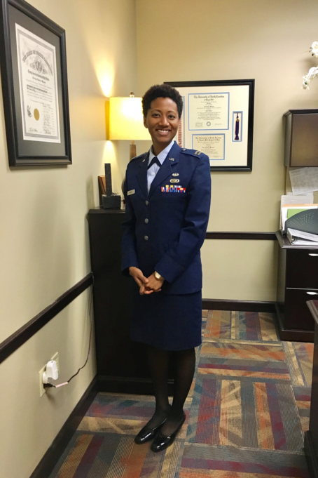 Mallette is photographed in her JAG uniform, a navy blue suit with a pale blue shirt. She stands in a brightly lit office and is smiling.