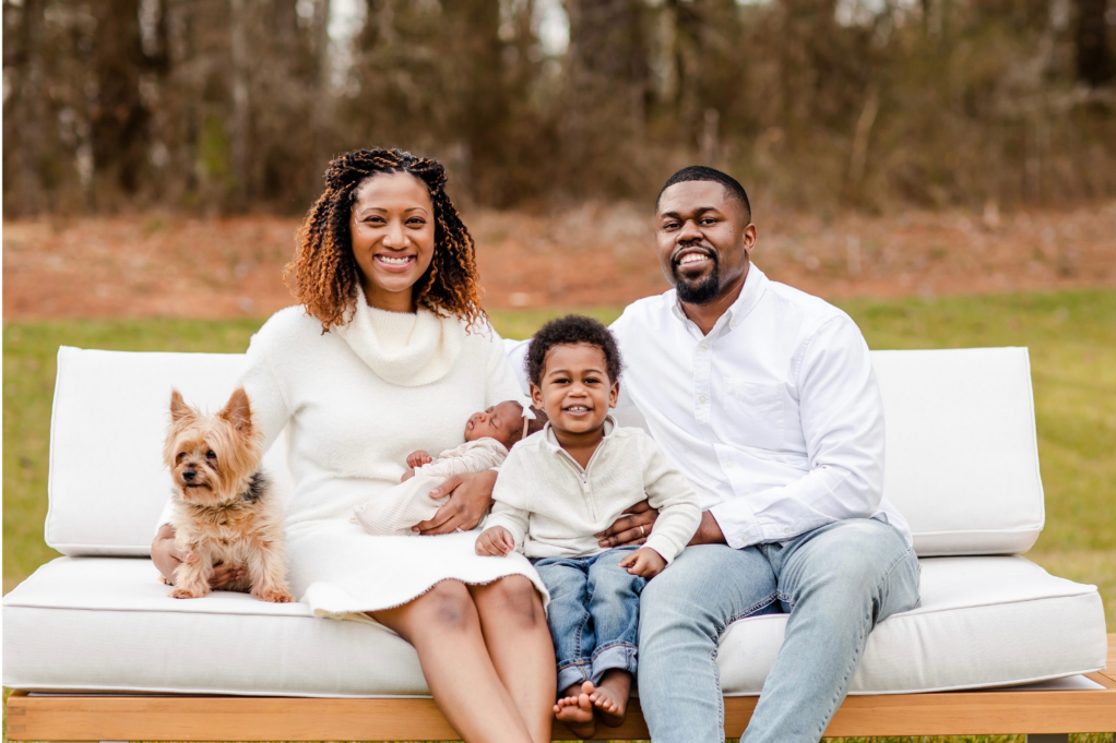 Mallette wears an ivory sweater dress and sits on a couch. Her husband, son, and pet dog sit next to her, and she holds her baby daughter in her arms.