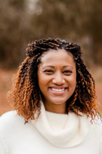 Jocelyn, a Black woman with black hair and gold lowlights, wears a cream-colored sweater and is pictured smiling.