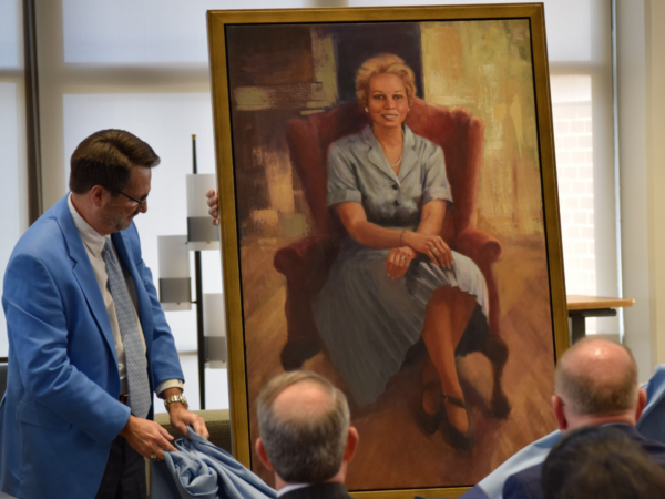 Judge Ammons stands to the left of the portrait of Sylvia, in which Sylvia, the subject, wears a blue dress and sits in a red chair.