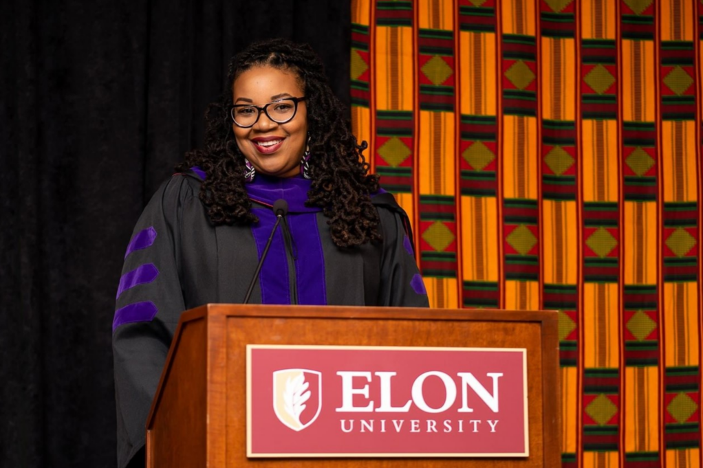 Tiffany, a Black woman, wears her law school graduation gown and stands in front of a podium that reads "Elon University."