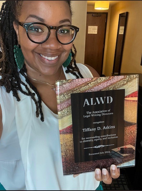 Tiffany, a Black woman with black hair, is wearing glasses and a white shirt. She holds the Association of Legal Writing Directors award, which is black with a sandy patterned frame.