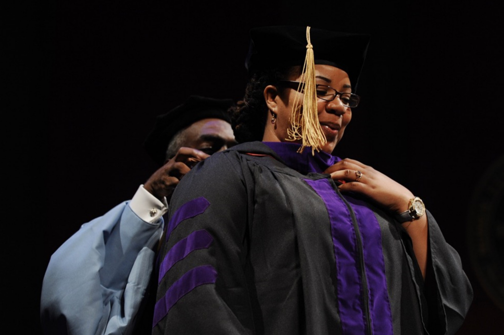 Tiffany, a Black woman, wears a graduation cap and gown with purple detailing. The hood is being placed on her by a Black man.