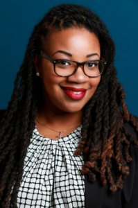 Tiffany, a Black woman with black hair and glasses, wears a white and black patterned shirt and black jacket and is pictured smiling.