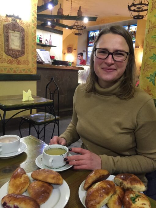 Svitlana is a young woman with brown hair and brown glasses. She wears an olive turtleneck and is pictured drinking a cup of coffee in a cafe.
