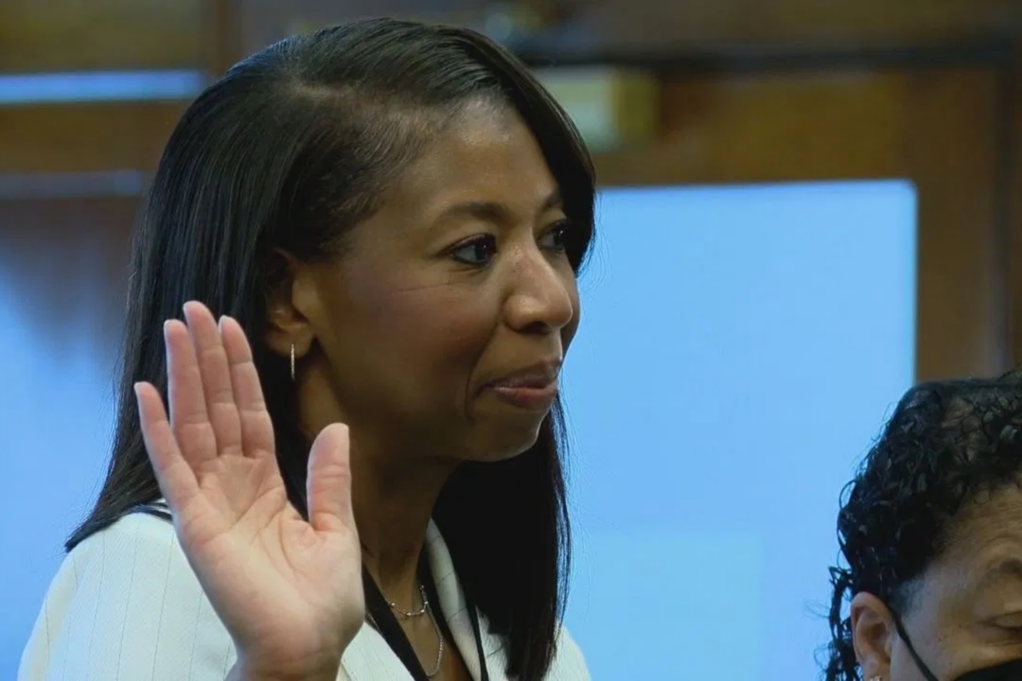 Jackie, a Black woman with black hair, is pictured wearing an ivory suit and is raising her right hand while being sworn in.