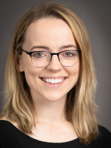 Katie, a woman with blond hair, wears black glasses and a black blouse. 