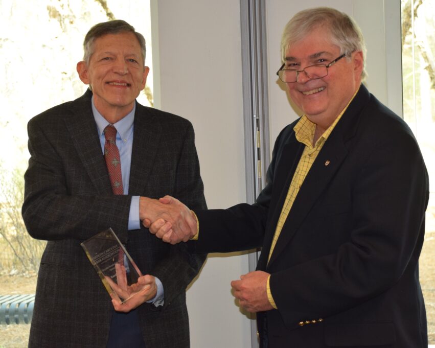 Judge Ward is a white man with grey hair and he wears a light blue shirt, red tie, an dark blue suit. Judge Ward holds his award and is shaking hands with Frank. 