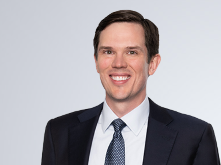 Erik, a man with dark brown hair, wears a white shirt, grey tie, and black jacket.