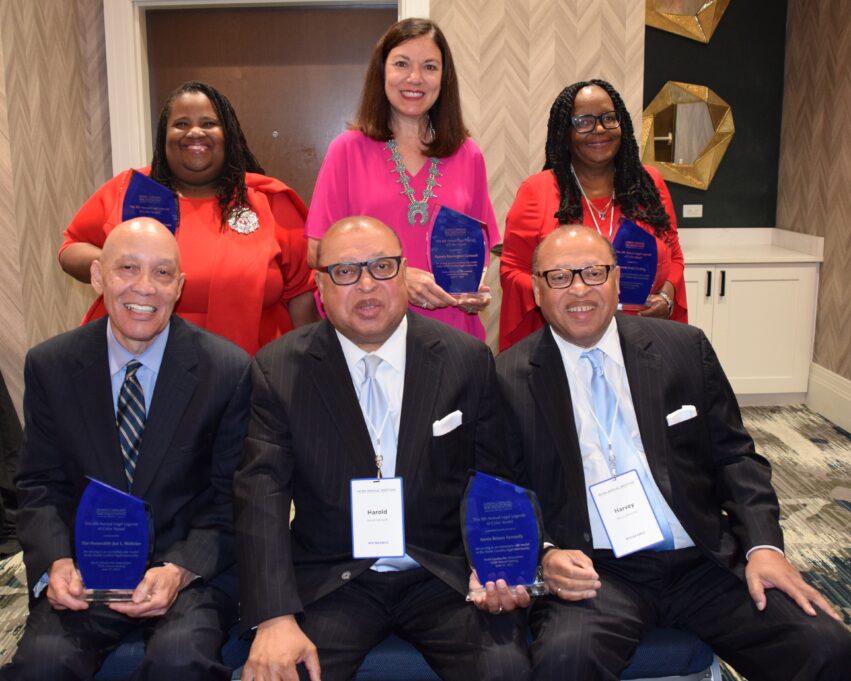 Judge Webster, a Black man, wears a blue shirt, navy tie, and black suit, Harold, a Black man, wears a white shirt, grey tie and black suit, Harvey Kennedy, a Black man, wears a white shirt, pale blue shirt, and black stripe suit, Judge Rawls, a Black woman with black hair, wears a red dress, Pamela, an American Indian woman with brown hair, wears a bright pink blouse, and Harding, a Black woman with black hair, wears a red dress.