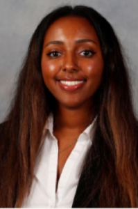 Iman, a woman with dark brown hair, wears a white shirt and black jacket.