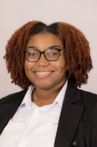 Jordyn, a Black person with short dark auburn hair, wears brown glasses, a white shirt and black jacket.