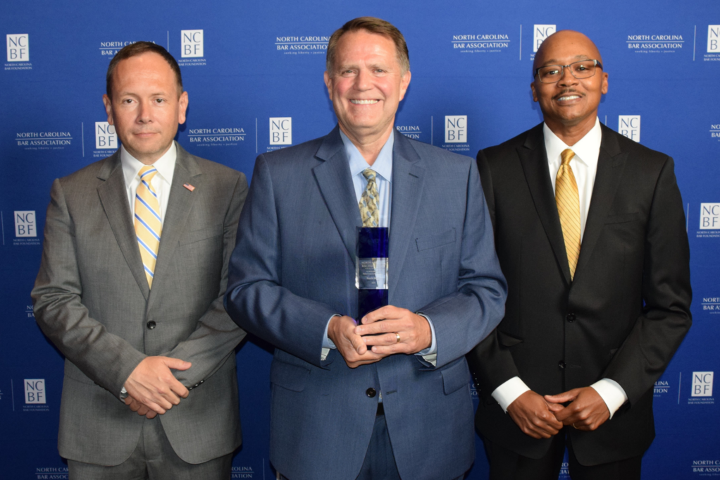 Gantt, a white man with brown hair, wears a pale blue shirt, grey suit, and off-white and pale blue tie and is holding the award. Jason is to his left and Clayton is to his right.