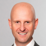 Cary, a white man with a shaved head, wears a white shirt, purple tie, and grey jacket. 