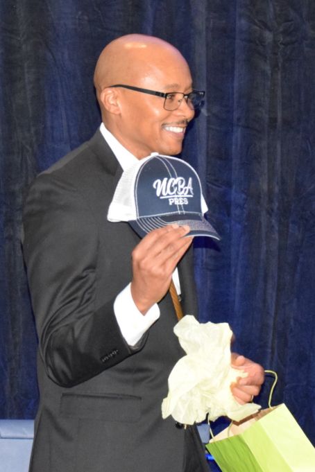 Clayton, a Black man with a shaved head, wears a white shirt and black suit and is pictured smiling and holding a baseball had that reads "NCBA pres." He has just opened a gift bag containing the hat.