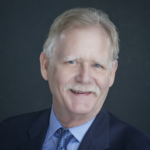 Fred, a white man with grey hair, wears a blue shirt, dark blue tie, and black jacket. 