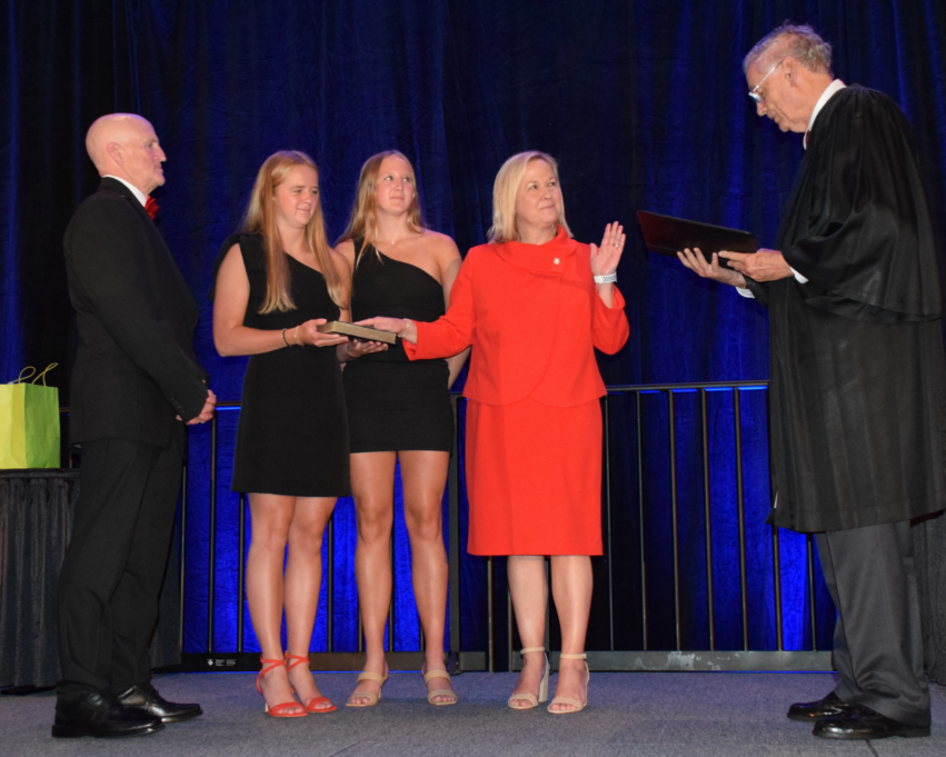 Patti, a white woman with blond hair, weras a red suit jacket and skirt. Her daughters, dressed in black dresses, stand to her left, and her spouse Chris is on the far left dressed in a suit. Judge Walker stands to the far right.