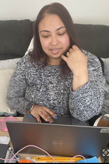 Elysia, a woman with dark brown hair, wears a grey sweater. She sits on a couch with her computer open and her phone on the table in front of her. She is volunteering from home as a part of NCBA 4ALL Statewide Day of Service.