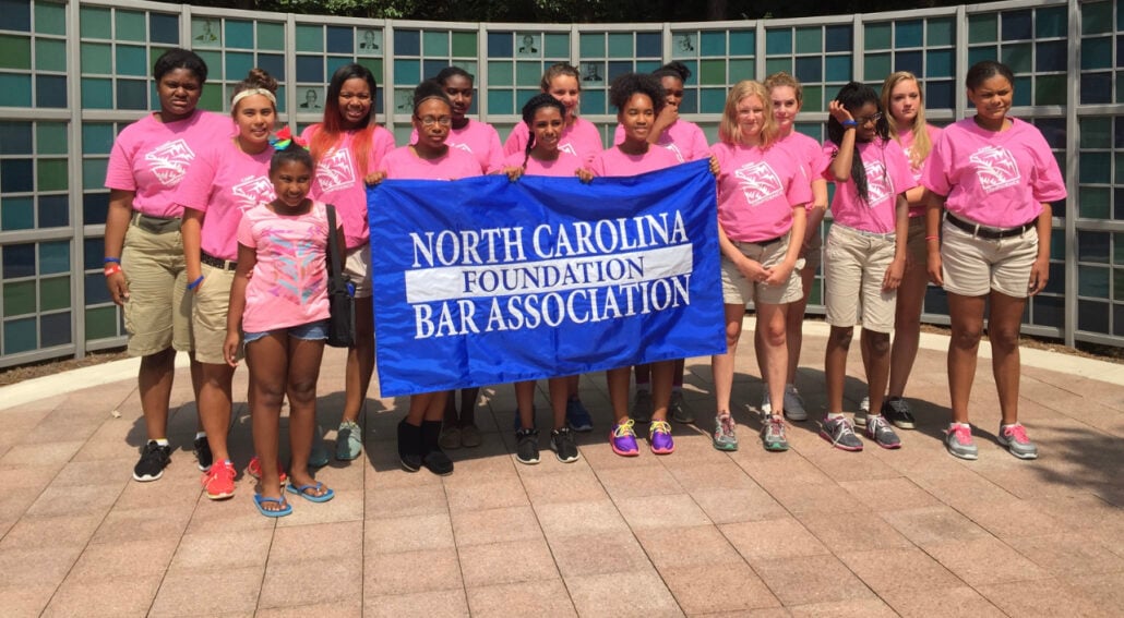 A photograph of Camp Confidence, an NCBA event, shows fourteen students dressed in pink shirts. The students hold a blue sign that reads "North Carolina Foundation Bar Association" in white. Elysia's daughter Saliyah stands on the far left.