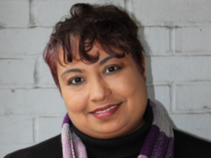 Elysia Prendergast-Jones, a woman with short brown hair, wears a black turtleneck and a purple scarf with lavender and dark purple stripes. She is smiling.