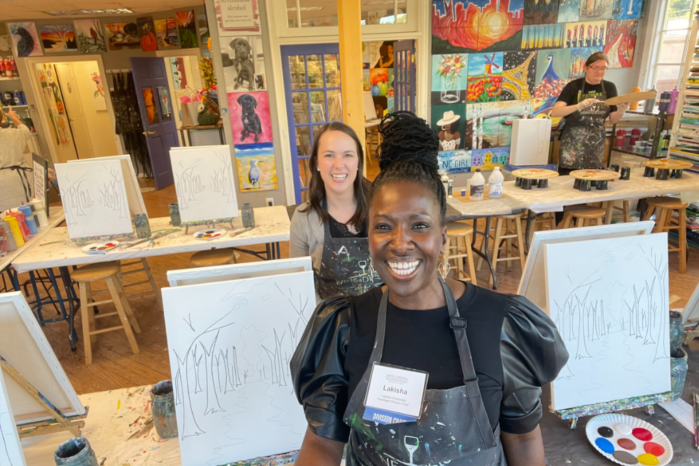 Lakisha, a Black woman with Black hair, stands smiling in front of an easel at the Wine & Design social.