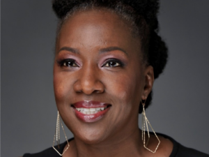 Lakisha Chichester, a Black woman with black hair, wears a black blouse and gold earrings. She is smiling.