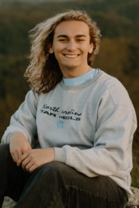 Max Macchia, a white young man with wavy blond hair, wears a grey sweatshirt that says "NC Tar Heels" in black. He sits on a mountaintop.