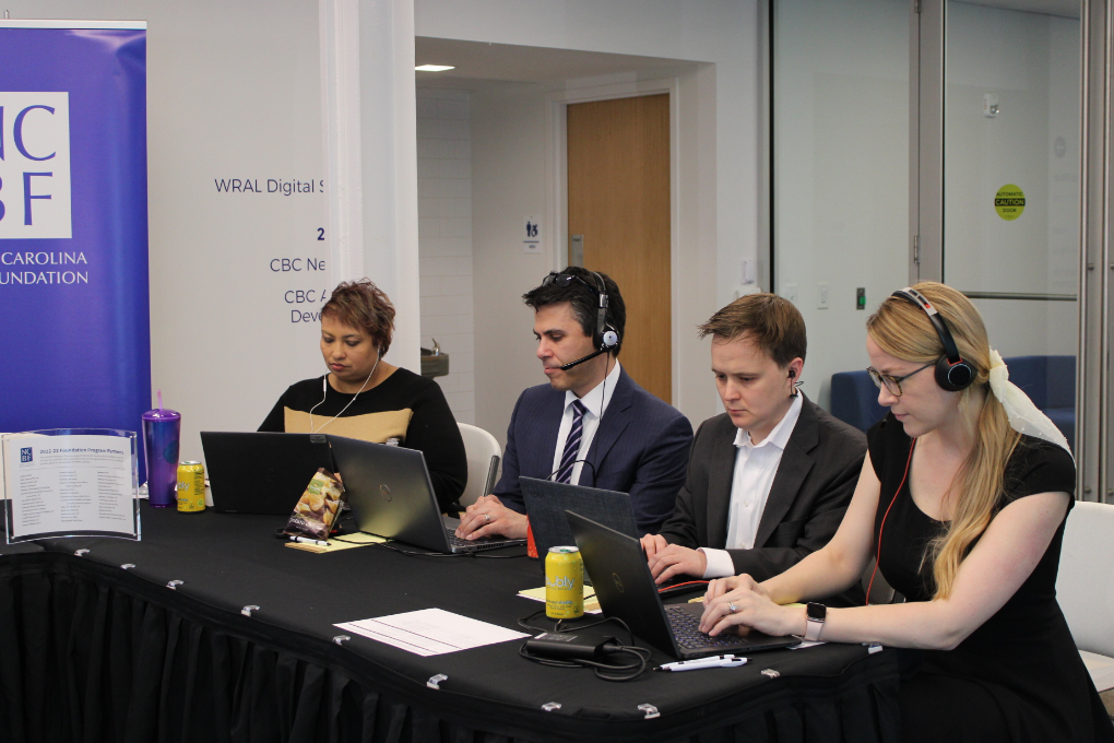 Elysia, a woman with short brown hair, wears a black and taupe long-sleeve dress. She sits at a computer with headphones on and is at a table with three other volunteers. 