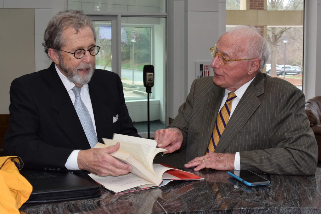 Cheshire, a white man with grey hair and a beard, wears black round glasses, a white shirt, grey tie and a black suit. Wade Smith, a white man with white hair, wears brown glasses, a white shirt, a tan and navy striped tie, and a grey pinstripe suit. They are talking together and seated at a desk, where Wade points to a book that Cheshire holds open.