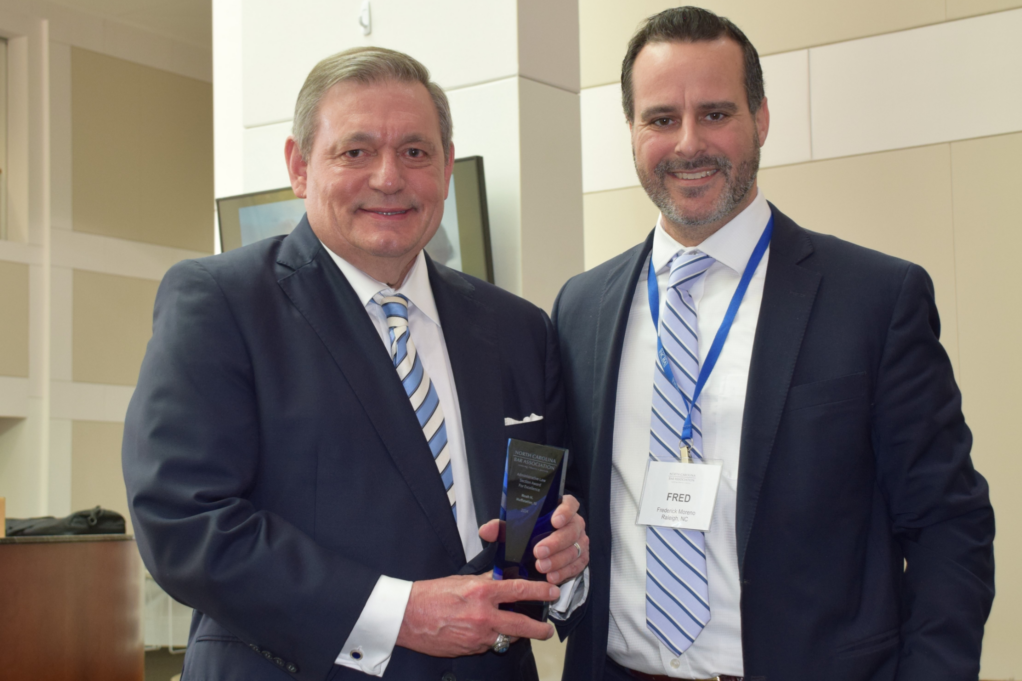 Noah, a white man with light brown hair, wears a white shirt, blue and white-striped tie, and dark grey suit. He holds a small blue award and stands with Fred Moreno, a man with brown hair and a beard, a white shirt, a pale blue and dark blue-striped tie and a black suit.