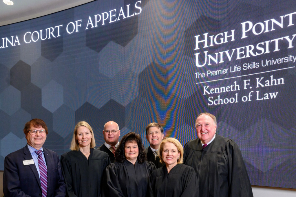 Mark Martin, a white man with brown hair and glasses, wears a pale blue shirt, navy suit, and purple and blue-striped tie. He stands with the N.C. Court of Appeals judges, who are dressed in robes, and the backdrop behind them reads, "Court of Appeals" and "High Point University, The Premier Life Skills University, Kenneth F. Kahn School of Law."