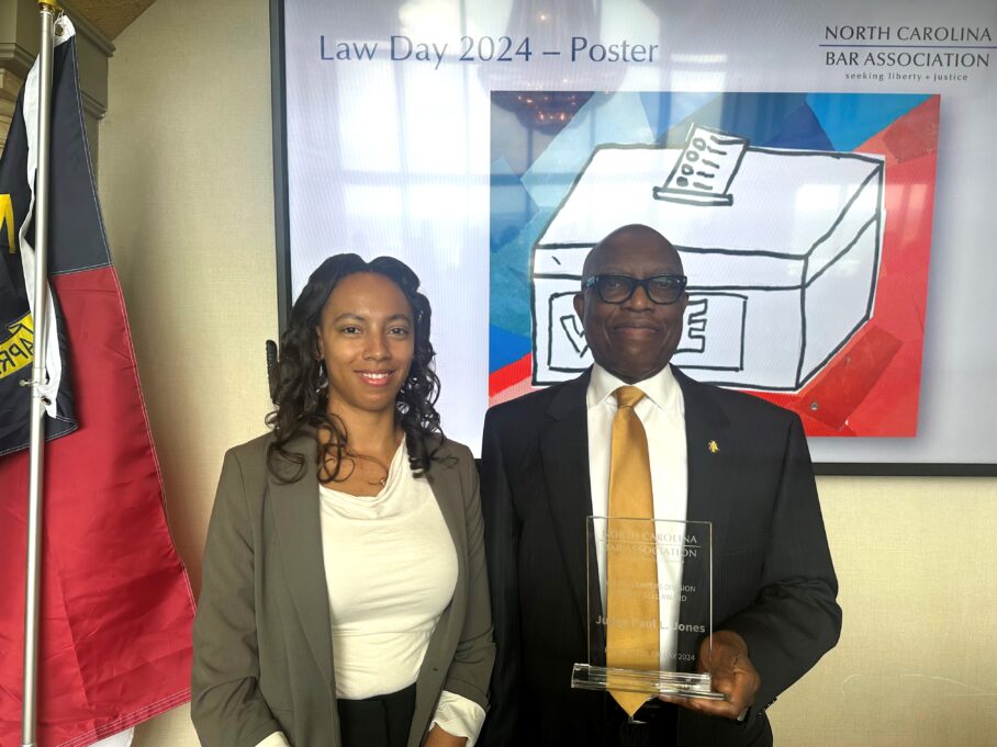 Paul Jones, a Black man with black glasses, wears a white shirt, gold tie and black jacket with a small black pin on the lapel. He stands with Sidney, a Black woman with curly black hair who wears a white shirt, black pants and light grey jacket. Behind them is a screen where a Law Day 2024 Poster appears with a red, white and blue image of a ballot box.