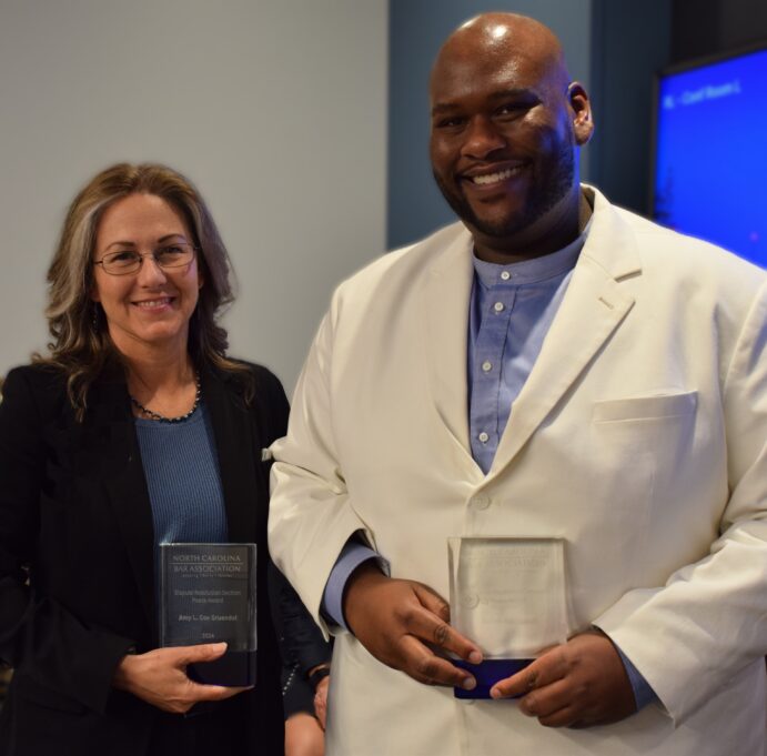 Amy, a white woman with curly brown hair and brown glasses, wears a blue shirt and black jacket and holds a clear glass award. Salim, a Black man with a beard, wears a blue shirt and a white jacket, and he also holds a clear glass award.