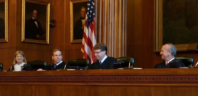 Mark Martin, a white man with brown hair and brown glasses, wears white shirt, blue tie and judicial robe. He sits on the bench with Paul Newby to his right and Robin Hudson and Robert Edmunds to his left. The American flag is behind him.