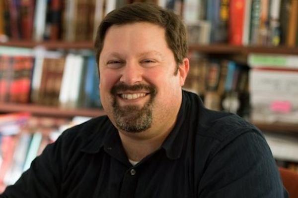 David Levine, a white man with brown hair and a beard, wears a black button down shirt. Books on a bookshelf are in the background.