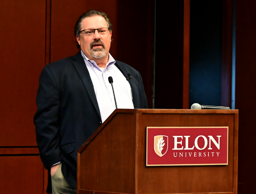 David, a white man with brown hair and a beard, wears a white shirt and black jacket. He stands speaking at a podium that has the Elon logo on the front. The logo has a red background and white letters. 